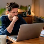 Homme assis devant ordinateur portable et factures empilees